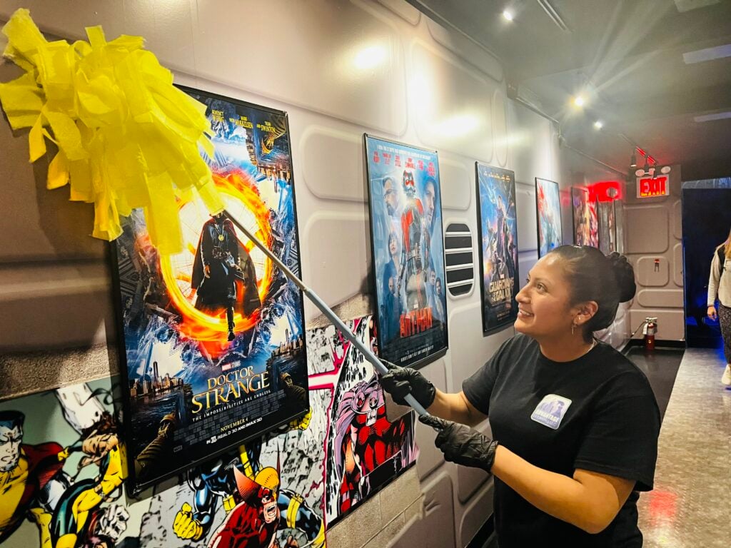 An Advantage Cleaning LLC worker dusting movie posters in a theater hallway decorated with Marvel superhero artwork.